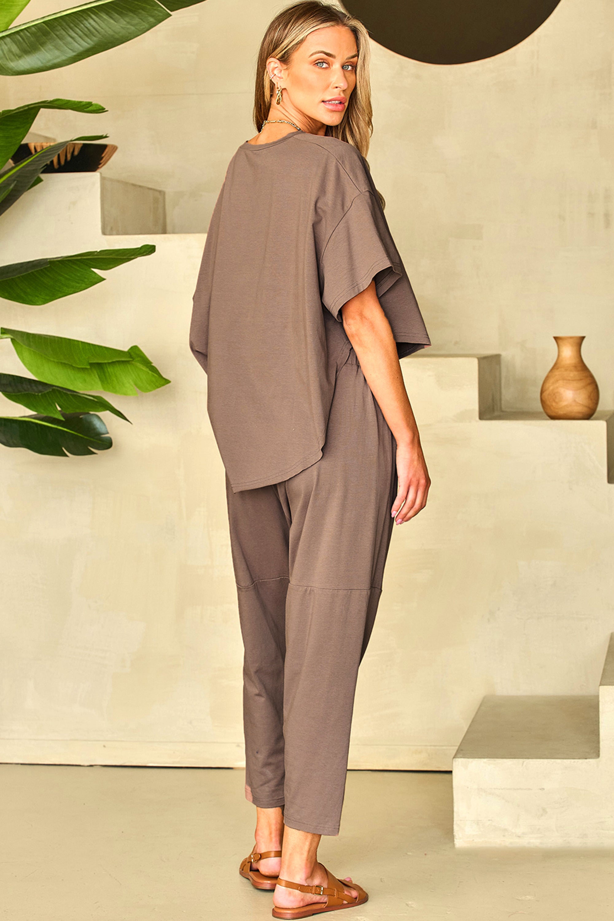 طقم نسائي مكون من بلوزة واسعة بأكتاف منسدلة قصيرة من الأمام وطويلة من الخلف، مع بنطلون بخصر قابل للتعديل وقصة مريحة وقصيرة. مصنوع من قماش ناعم ومرن، ومثالي للاسترخاء أو لزياراتك الغير رسمية. لون بني رمادي.